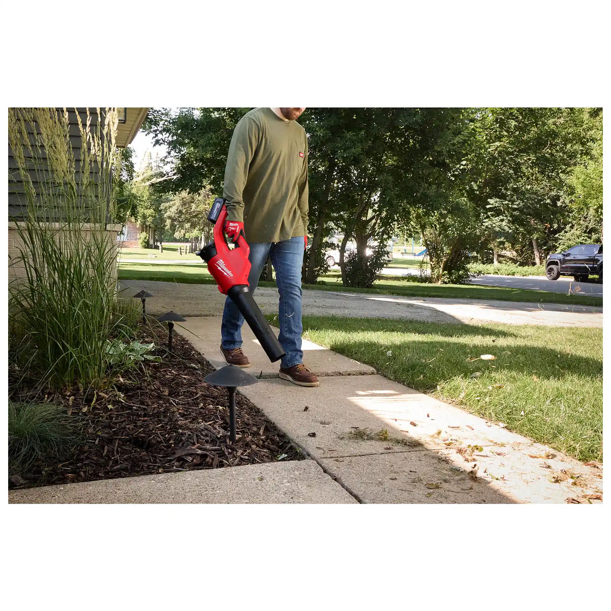 A person uses the M18™ Brushless Blower to clear leaves from a sidewalk adjacent to a landscaped garden. The blower is red and black with a comfortable grip handle. The scene takes place during the day in a suburban outdoor setting.