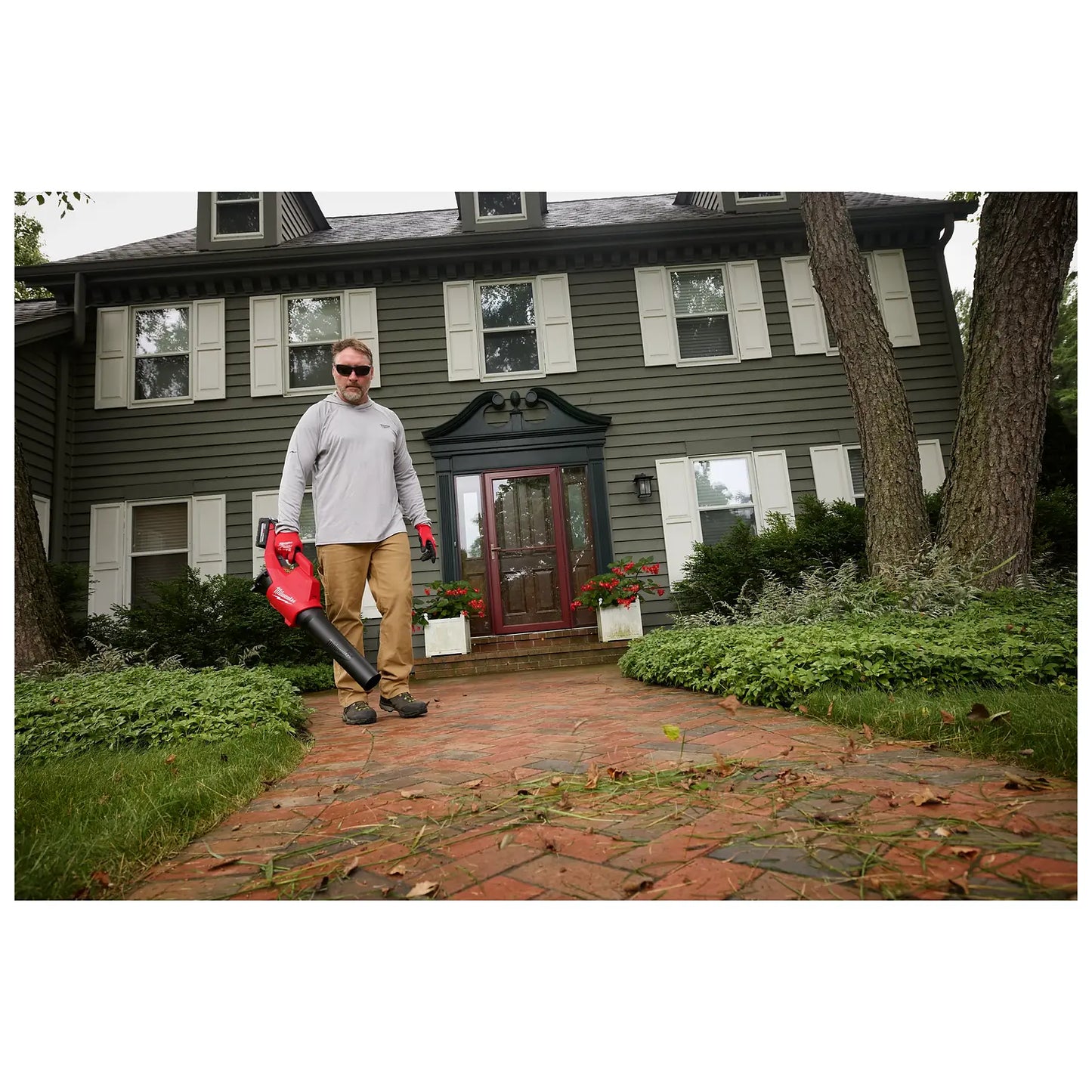 A person stands on a brick walkway in front of a green, two-story house holding an M18™ Brushless Blower. The blower is red with a black nozzle. Green bushes and trees surround the house, and fallen leaves are visible on the walkway.