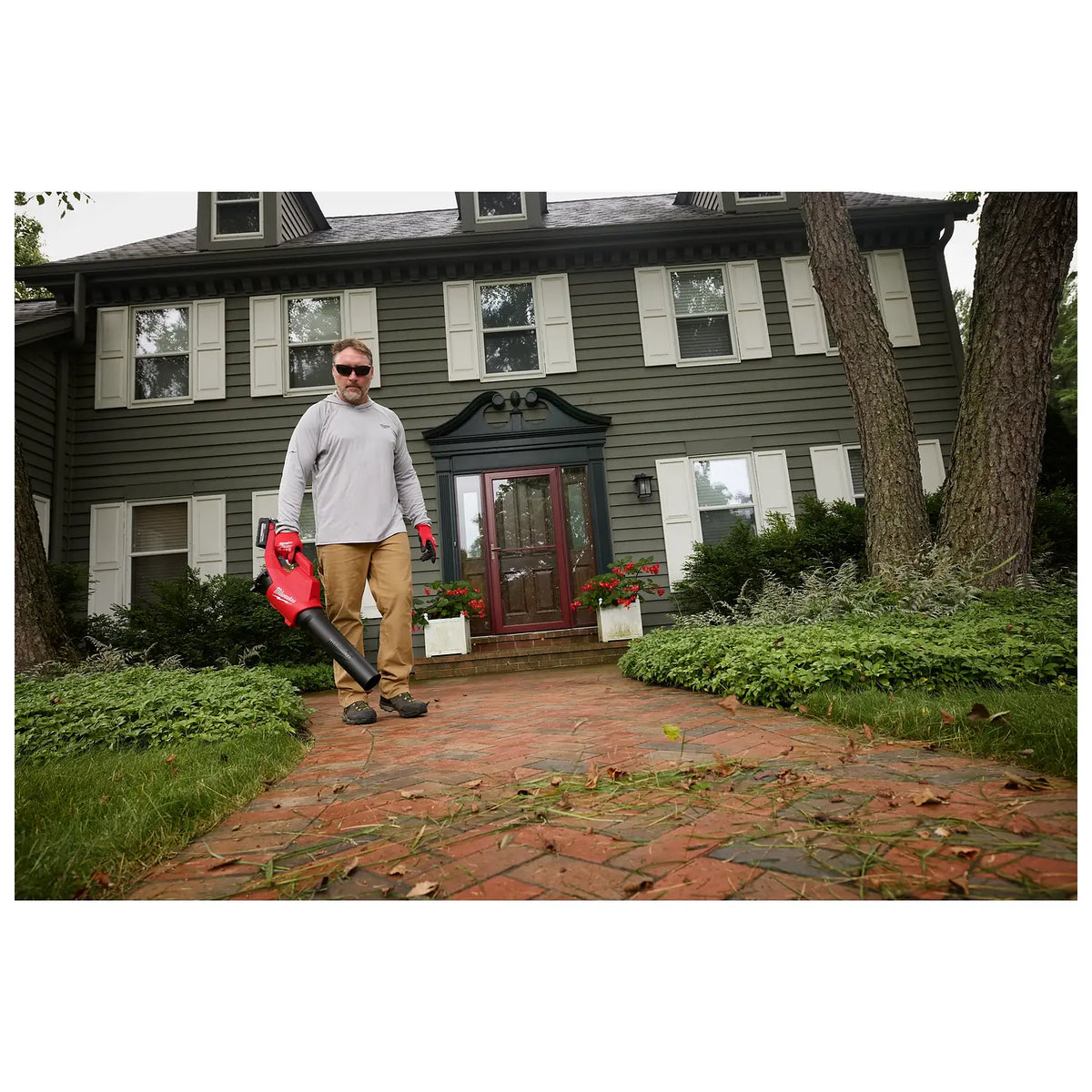 A person stands on a brick walkway in front of a green, two-story house holding an M18™ Brushless Blower. The blower is red with a black nozzle. Green bushes and trees surround the house, and fallen leaves are visible on the walkway.