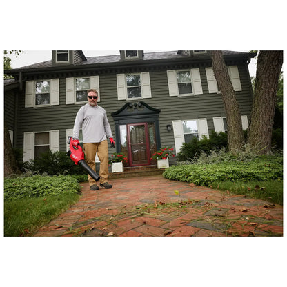 A person stands on a brick walkway in front of a green, two-story house holding an M18™ Brushless Blower. The blower is red with a black nozzle. Green bushes and trees surround the house, and fallen leaves are visible on the walkway.