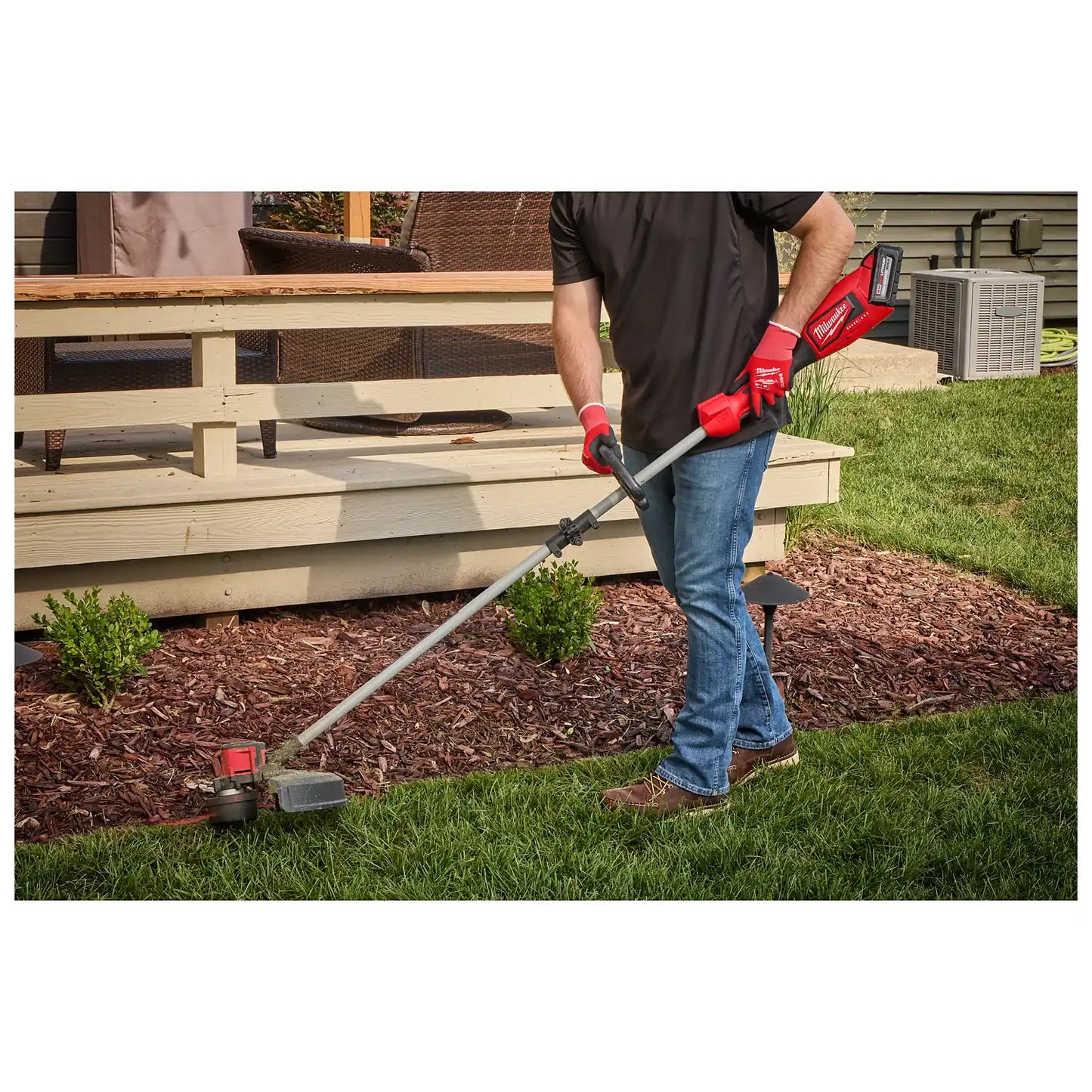 A person using the M18™ Brushless String Trimmer to trim grass along the edge of a garden bed. The trimmer has a red and black design with a long shaft and battery pack at the end. The user is wearing red gloves and jeans.
