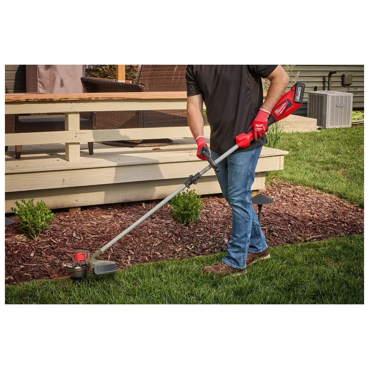 A person using the M18™ Brushless String Trimmer to trim grass along the edge of a garden bed. The trimmer has a red and black design with a long shaft and battery pack at the end. The user is wearing red gloves and jeans.