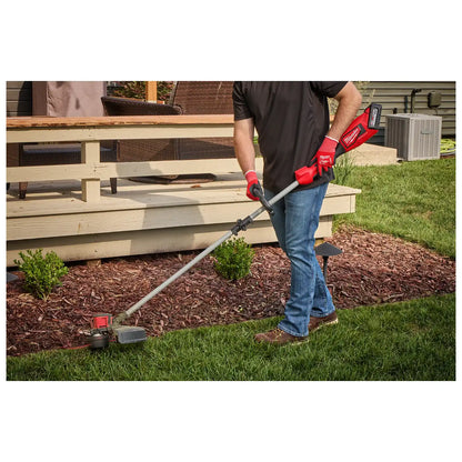 A person using the M18™ Brushless String Trimmer to trim grass along the edge of a garden bed. The trimmer has a red and black design with a long shaft and battery pack at the end. The user is wearing red gloves and jeans.