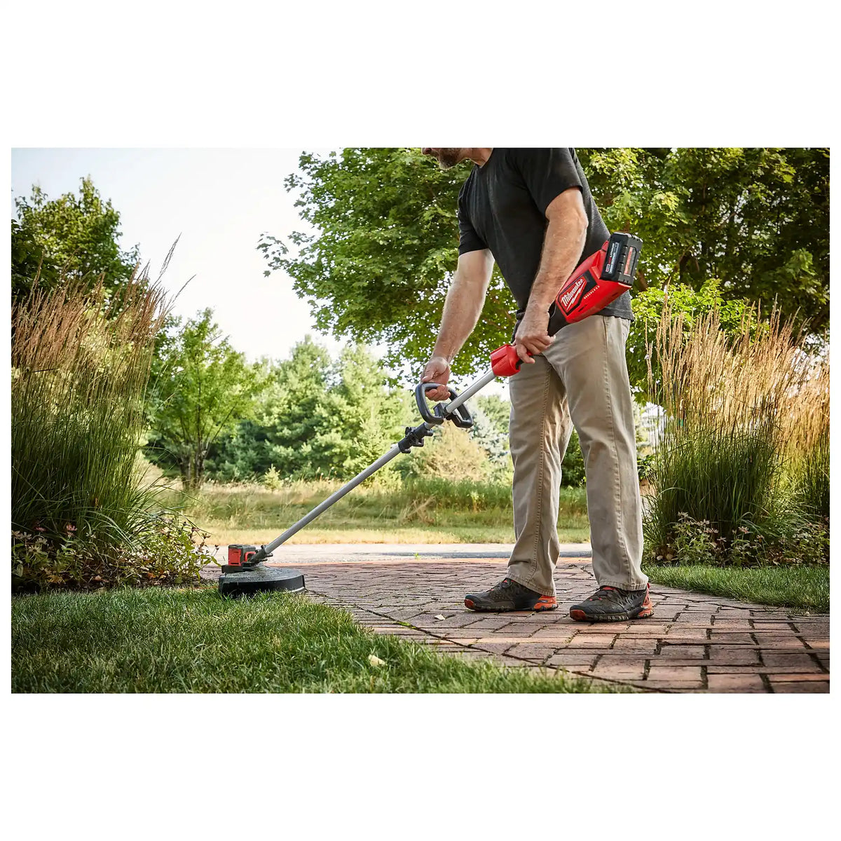 A man uses the M18™ Brushless String Trimmer to edge a lawn beside a brick walkway. The trimmer features a long handle for better reach and a red-colored motor casing, highlighting its battery-operated design. Dense greenery and tall grass surround the path in an outdoor setting.