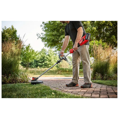 A man uses the M18™ Brushless String Trimmer to edge a lawn beside a brick walkway. The trimmer features a long handle for better reach and a red-colored motor casing, highlighting its battery-operated design. Dense greenery and tall grass surround the path in an outdoor setting.